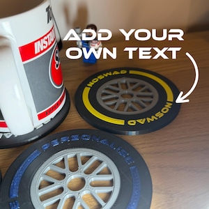 Könnte beinhalten: Zwei kreisförmige Untersetzer im Design von Rennreifen mit grauen Radspeichen. Ein Untersetzer hat ein gelbes Felgendetail mit dem Text NOSWAD, der andere blaue Schriftzüge mit der Aufschrift PERSONALISE. Ein weißer Becher und eine Spielfigur stehen daneben.