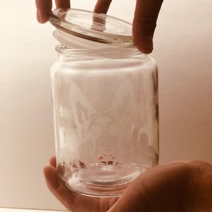 Pode incluir: Um frasco de vidro transparente com tampa, apresentando um desenho gravado de uma cabe&ccedil;a de c&atilde;o. O frasco est&aacute; sendo segurado por uma m&atilde;o.