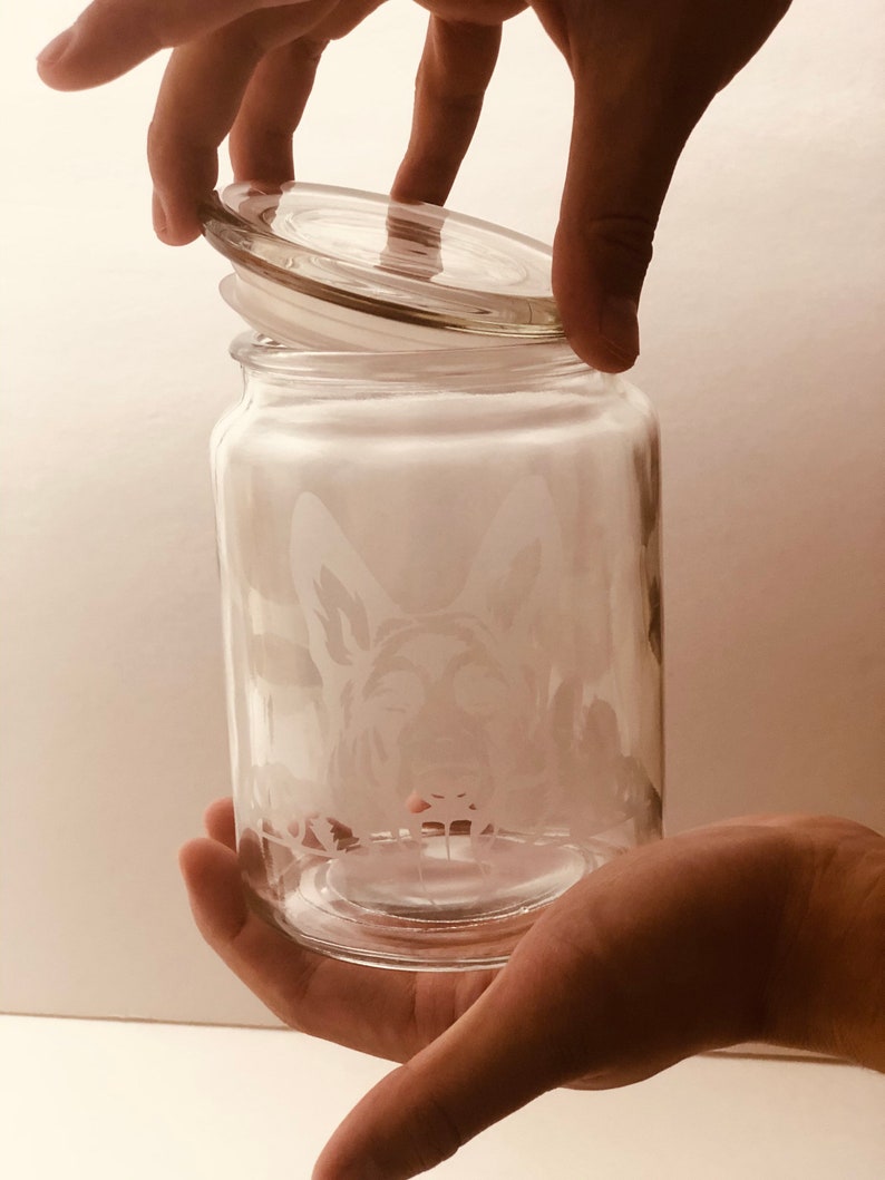 Pode incluir: Um frasco de vidro transparente com tampa, apresentando um desenho gravado de uma cabe&ccedil;a de c&atilde;o. O frasco est&aacute; sendo segurado por uma m&atilde;o.
