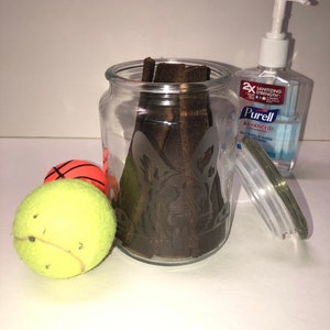 Pode incluir: Um frasco de vidro com uma silhueta de c&atilde;o gravada na lateral, cheio de mastig&aacute;veis para c&atilde;es marrons. Duas bolas de t&eacute;nis e uma bola de basquete est&atilde;o em primeiro plano. Uma garrafa de desinfetante para as m&atilde;os Purell Advanced est&aacute; no fundo.