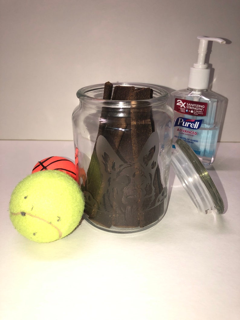 Pode incluir: Um frasco de vidro com uma silhueta de c&atilde;o gravada na lateral, cheio de mastig&aacute;veis para c&atilde;es marrons. Duas bolas de t&eacute;nis e uma bola de basquete est&atilde;o em primeiro plano. Uma garrafa de desinfetante para as m&atilde;os Purell Advanced est&aacute; no fundo.