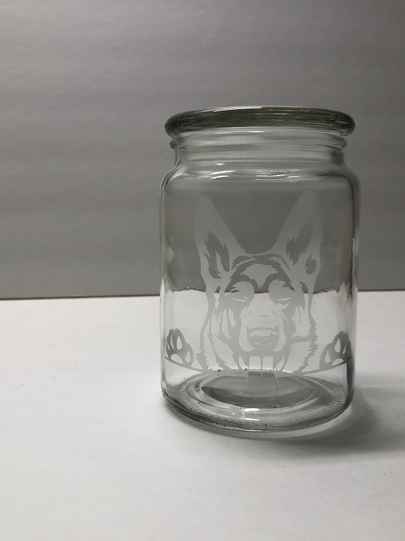 Pode incluir: Um frasco de vidro transparente com tampa, apresentando um design gravado da cabe&ccedil;a de um c&atilde;o pastor alem&atilde;o olhando por cima.