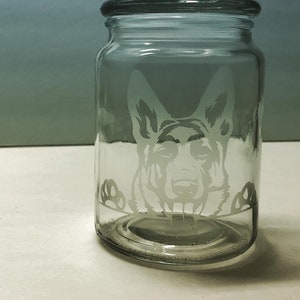 Pode incluir: Um frasco de vidro transparente com tampa, apresentando um design gravado da cabe&ccedil;a de um c&atilde;o pastor alem&atilde;o espiando pela borda.