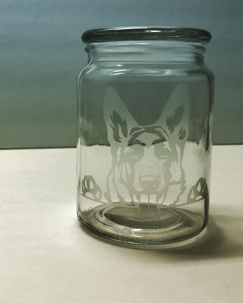Pode incluir: Um frasco de vidro transparente com tampa, apresentando um design gravado da cabe&ccedil;a de um c&atilde;o pastor alem&atilde;o espiando pela borda.