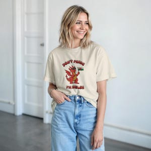 Puede incluir: Camiseta color crema con un dragón de dibujos animados sosteniendo una bandera galesa. El texto "DON'T PANIC I'M WELSH" está impreso en rojo y negro. La modelo lleva vaqueros azul claro.