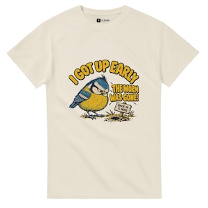 Może przedstawiać: Kremowy t-shirt z krótkim rękawem z grafiką przedstawiającą niebiesko-żółtego ptaka patrzącego na dziurę w ziemi. Tekst głosi I GOT UP EARLY THE WORM WAS GONE!, a mała tabliczka obok dziury zawiera napis BACK IN 5 MINS.