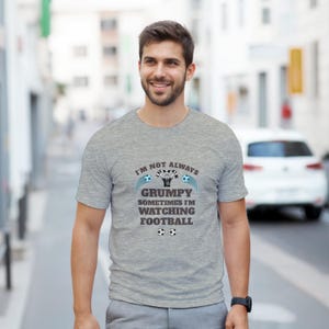 Op de afbeelding: Een lichtgrijs t-shirt met de tekst "I'M NOT ALWAYS GRUMPY SOMETIMES I'M WATCHING FOOTBALL" in zwart en blauw. Het shirt heeft voetbalafbeeldingen. De persoon draagt een zwart horloge.