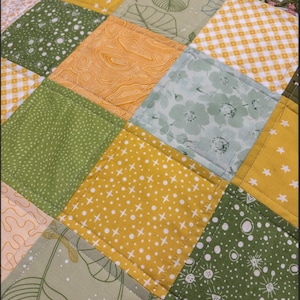 May include: Close-up of a patchwork quilt with a variety of square patterns. The squares feature floral, leaf, and geometric designs in shades of green, yellow, and white. The quilt is machine-stitched.