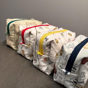 Peut inclure: Quatre trousses de maquillage matelassées avec des fermetures éclair de couleurs différentes : vert, rouge, jaune et bleu marine. Les sacs sont blancs avec un motif répété d'illustrations noires. Les sacs sont disposés en rangée sur une surface grise.