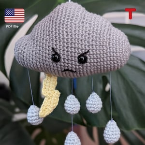 May include: A crocheted gray cloud with a yellow lightning bolt and blue raindrops hanging from it. The cloud has a grumpy face with black eyes and a frown.