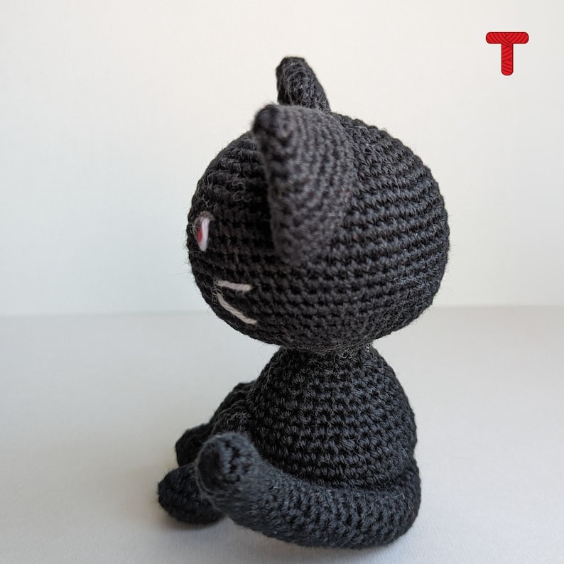 May include: A black crocheted cat toy with white embroidered facial features. The cat is sitting with its tail curled around its body.