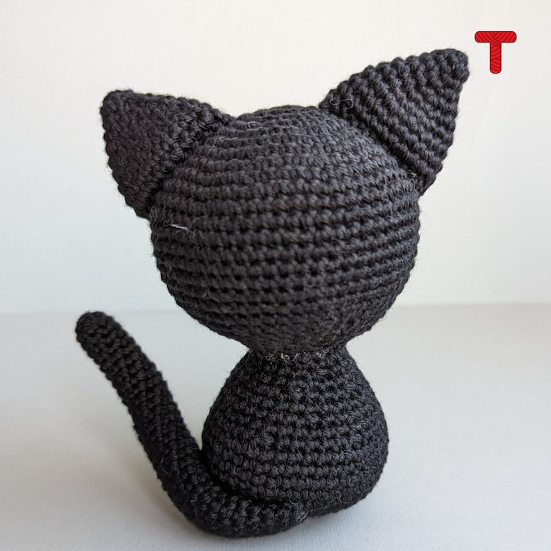 May include: A black crocheted cat toy with a long tail. The cat is sitting on a white surface.