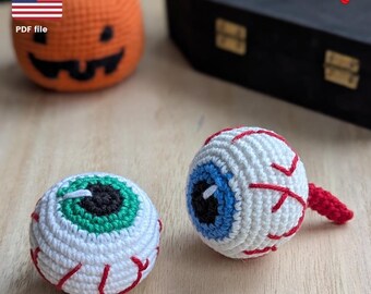 Könnte beinhalten: Gehäkelte Halloween-Dekorationen, darunter zwei Augapfel-Ornamente mit roten Adern, weißer Lederhaut und blauen und grünen Iris. Ein orangefarbener Kürbis und ein schwarzer Sarg sind im Hintergrund. Enthält ein PDF-Dateisymbol und die amerikanische Flagge.