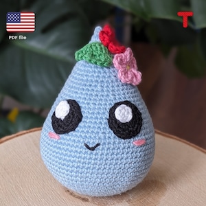 May include: A blue crocheted raindrop-shaped plush toy with a smiling face, big eyes, and a flower crown. The crown is made of green, red, and pink crocheted flowers.