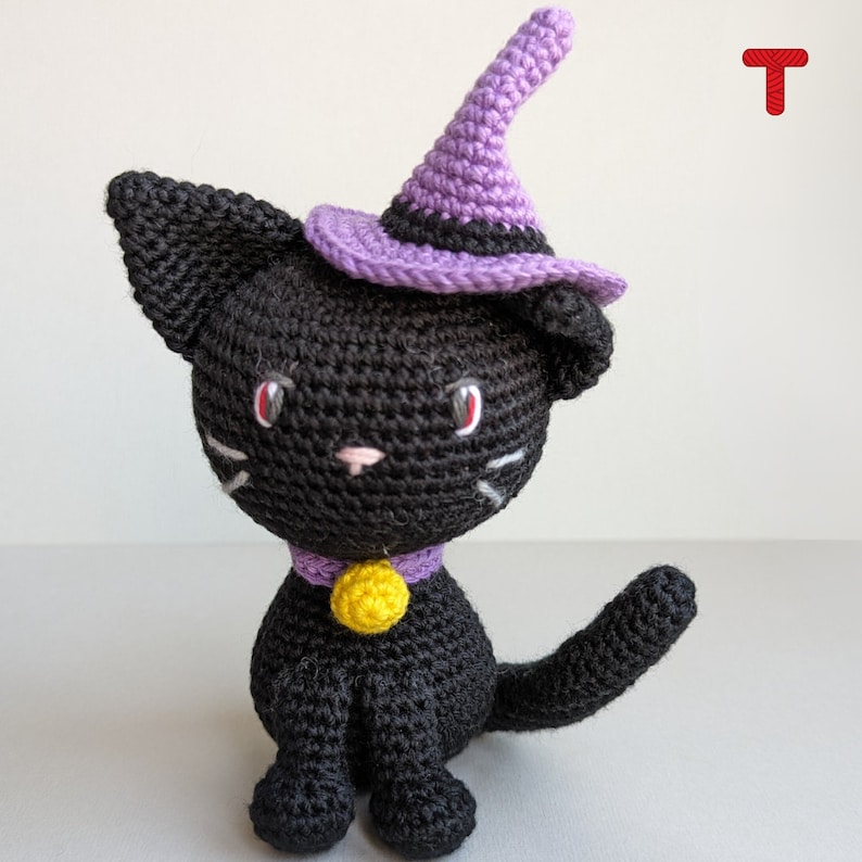 May include: A black crocheted cat wearing a purple witch hat with a yellow bell around its neck.