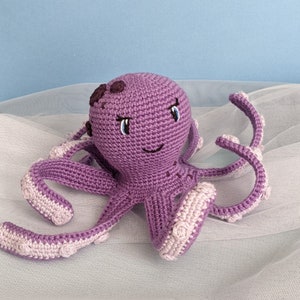 May include: A purple crocheted octopus with embroidered eyes and a smiling mouth. The octopus has eight tentacles with white accents.