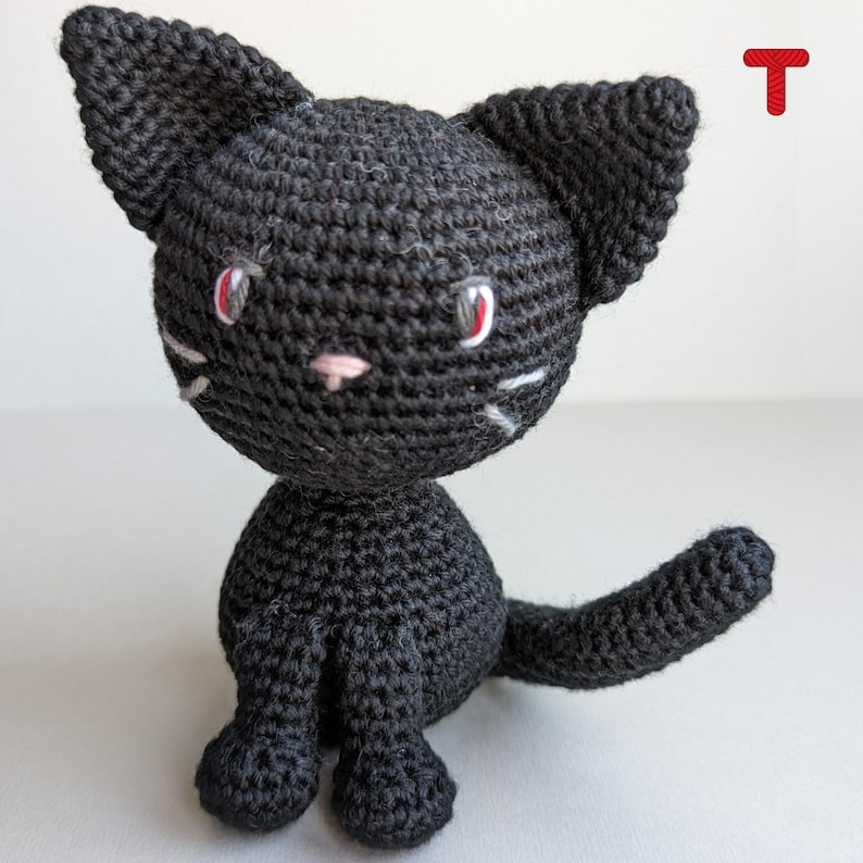 May include: A black crocheted cat with white whiskers and red eyes. The cat is sitting with its tail curled behind it.