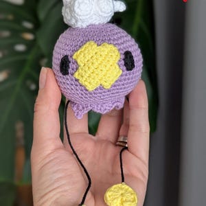 May include: A handmade crochet plush toy in the shape of a purple creature with a yellow face, black eyes, and a white cloud on top. It has two black dangling strings with yellow flower-shaped accents at the ends.