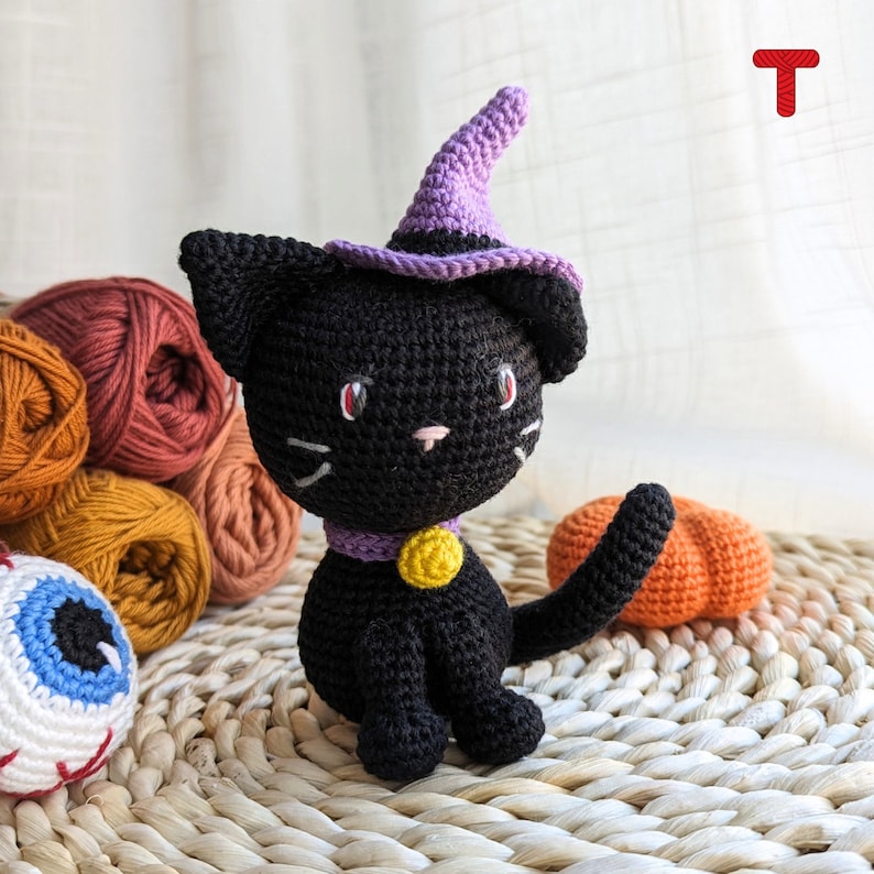 May include: A black crocheted cat wearing a purple witch hat with a yellow bell around its neck. The cat is sitting on a woven wicker surface with a small orange crocheted pumpkin beside it.
