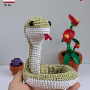 May include: A crocheted green and white snake toy with black eyes and a red tongue. The snake is curled up in a circle and is being held in a person's hand.