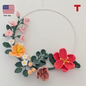 Puede incluir: Una corona floral de ganchillo con flores rosas, naranjas y rojas, hojas verdes y bayas blancas. La corona está colgada de un aro de metal blanco.