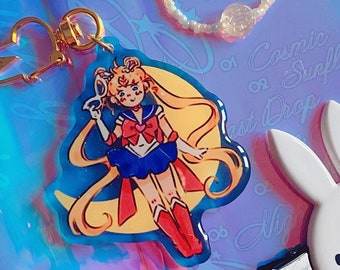 May include: A clear plastic bag with a variety of items inside, including a white beaded necklace, a small white bunny keychain, a charm with a cartoon character, and a blue and white patterned card with the text "Cosmic or Sunflower. Last Drop Reads".