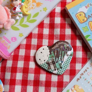 以下が含まれることがあります： 白、緑のチェック柄、チョコレートソースがかかった茶色のパーツを組み合わせたコラージュデザインのハート型ピンバッジ。ピンバッジには筆記体で「mini choco」という文字が記されており、周囲には様々な文房具が配置されています。