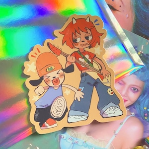 Könnte beinhalten: Ein Aufkleber mit zwei Comicfiguren. Eine ist eine rothaarige Figur mit einer Gitarre, die ein blaues Hemd und Jeans trägt. Die andere ist eine hundeartige Figur mit orange-weißem Fell, die ein blaues Hemd und Jeans trägt. Der Aufkleber hat einen bunten, regenbogenartigen Hintergrund.