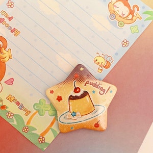 Könnte beinhalten: Ein sternförmiger Ansteck-Pin mit einem Pudding-Dessert mit Kirsche. Der Pin hat ein gelbes und braunes Farbschema mit dem Wort "pudding!" darauf. Der Pin befindet sich auf einem farbenfrohen Hintergrund mit Cartoon-Affen- und Bienen-Illustrationen.