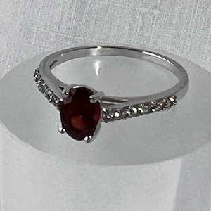 May include: A silver ring featuring a deep red oval gemstone. The band is embellished with small, clear stones on either side of the central gem. The ring is presented on a clear, cylindrical stand against a white backdrop.
