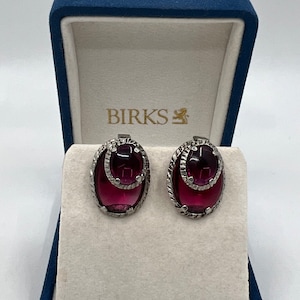 Op de afbeelding: Een paar zilveren oorbellen met een donkerpaarse, ovale steen die in een zilveren frame is gezet. De oorbellen zitten in een blauwe fluwelen doos met het woord "BIRKS" erop gedrukt.