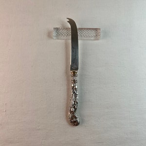 May include: A silver cheese knife with an ornate, detailed handle. The knife has a curved blade and is resting on a clear glass stand.