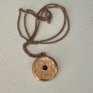 May include: A gold-toned metal locket necklace featuring a circular pendant with an intricate engraved floral scrollwork pattern and a small, centred dark red gemstone. The pendant hangs from a delicate, matching gold-toned metal chain link.