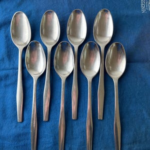 May include: Eight stainless steel spoons are displayed on a blue cloth. The spoons have a classic design with oval bowls and slim handles. The spoons are shiny, and the cloth has a textured appearance. The spoons are likely for dining.