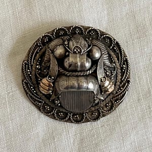 May include: An oval silver brooch with detailed metalwork. The central design features a stylized face with gold-toned accents. The brooch is surrounded by a rope-like border. The overall design is ornate and decorative.
