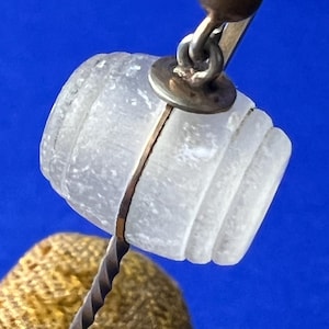 May include: A clear, barrel-shaped bottle stopper with a silver metal band and a twisted metal stem. The stopper has a small, gold-colored ball at the top and is set against a blue background.
