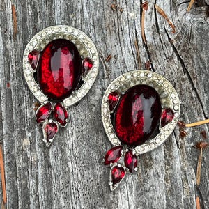 Peut inclure: Paire de boucles d'oreilles vintage à clip. Chaque boucle d'oreille présente une grosse pierre ovale rouge foncé entourée d'un cadre argenté orné de strass transparents. De plus petites pierres rouges sont placées en dessous et sur les côtés.