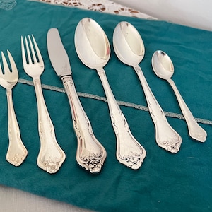 Könnte beinhalten: Ein Set aus sechs versilberten Besteckteilen, darunter ein Messer, eine Gabel und vier Löffel, mit verzierten Blumendesigns an den Griffen. Das Besteck ist auf einer tealfarbenen Stoffoberfläche angeordnet.