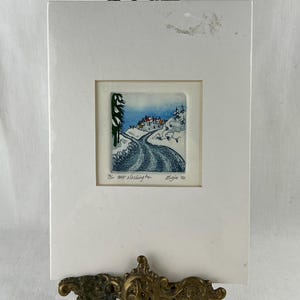 May include: A framed winter landscape art print. The artwork shows a snow-covered road leading to a village with red-roofed houses. The print is set in a white mount and displayed on a decorative brass easel. The text "The Mt. Washington" and "Zejia '92" are visible.
