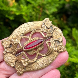 May include: An antique brooch with a gold-coloured metal frame. The brooch has floral and swirling patterns, and a central oval-shaped, deep red stone. The pin is held in a hand against a blurred green background.