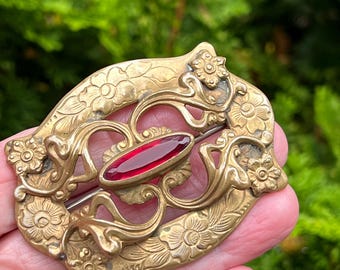 Op de afbeelding: Oud broche met een goudkleurig metalen frame. De broche heeft bloemen- en wervelende patronen en een centrale ovaalvormige, dieprode steen. De speld wordt in een hand gehouden tegen een vage groene achtergrond.