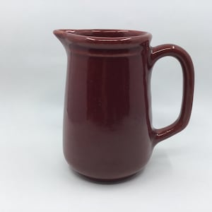 May include: A dark red ceramic jug with a handle. The jug is made of a smooth, glossy material.
