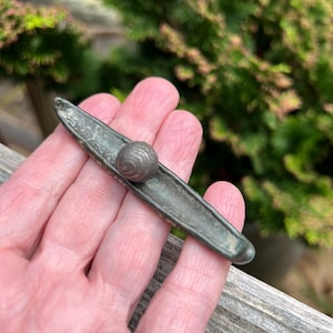 May include: A small, antique metal object with a long, narrow, boat-like shape and a spherical ball at the center. The metal has a weathered, aged appearance with a dark, greenish-gray patina. The object is held in a hand against a blurred green background.