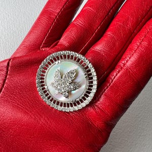 Peut inclure: Broche ronde argentée avec un centre en nacre et un motif de feuille d'érable étincelant. La broche est entourée d'un anneau de petits cristaux transparents. La broche est posée sur un fond en cuir rouge.