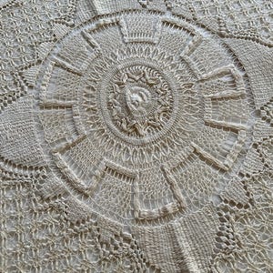 May include: Close-up of a cream-colored, intricately crocheted tablecloth. The design features a central circular motif with radiating patterns and geometric shapes. The detailed craftsmanship is evident in the delicate openwork and textured stitches.