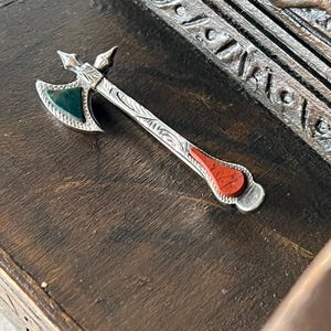 Puede incluir: Colgante de hacha en miniatura de color plateado con un mango detallado y detalles decorativos. La cabeza del hacha presenta una piedra verde en un lado y una piedra roja en el otro. El colgante se muestra sobre una superficie de madera oscura.