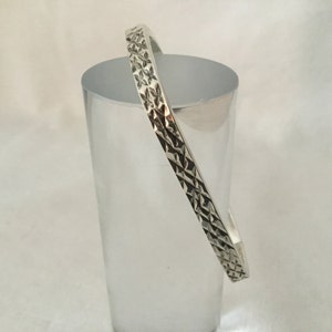 May include: A silver bangle with a geometric design. The bracelet, made of shiny metal, has a square profile and a repeating pattern of small shapes, creating a textured surface. It is shown on a clear, cylindrical object. The bangle would fit a wrist of approximately 17 cm.