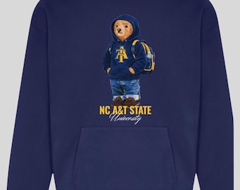 May include: Navy blue hoodie with a cartoon bear wearing a hoodie with the NC A&T State University logo. The bear is also wearing denim shorts and a backpack. The text "NC A&T STATE University" is printed below the bear.