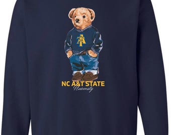 May include: Navy blue sweatshirt with a teddy bear illustration. The bear wears a blue sweater with a yellow and blue logo and blue jeans. The text "NC A&T STATE University" is printed below the bear.