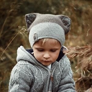 Puede incluir: Un niño pequeño con un suéter de punto gris y un gorro de punto gris y marrón con orejas de pelo. El gorro tiene una banda marrón alrededor de la corona.
